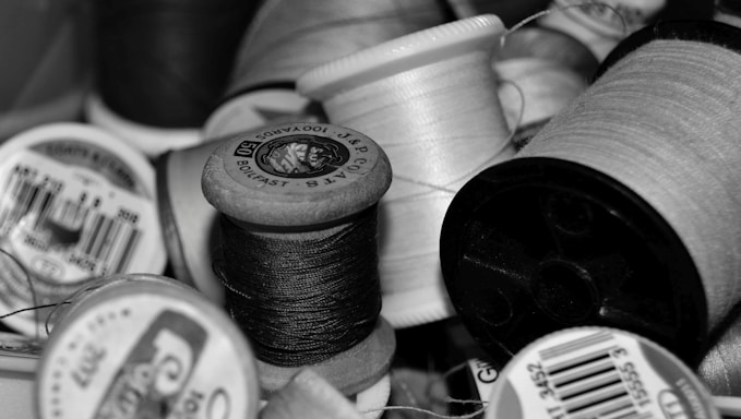 A collection of thread spools in various sizes and colors, close-up. The threads are tightly wound around the spools, and some have labels with brand names and product details. The arrangement appears random, showing a variety of textures and materials.