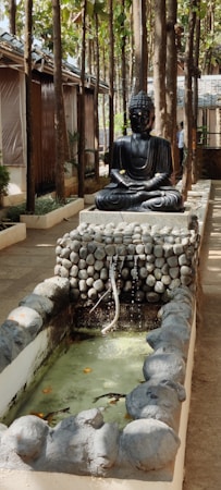 A serene outdoor setting with a statue of Buddha sitting in a meditative pose. The statue is black and surrounded by a backdrop of trees and rustic wooden structures. In front of the statue is a small water feature with stones, where water gently flows into a basin below. The environment has a tranquil and peaceful atmosphere with natural elements and greenery.