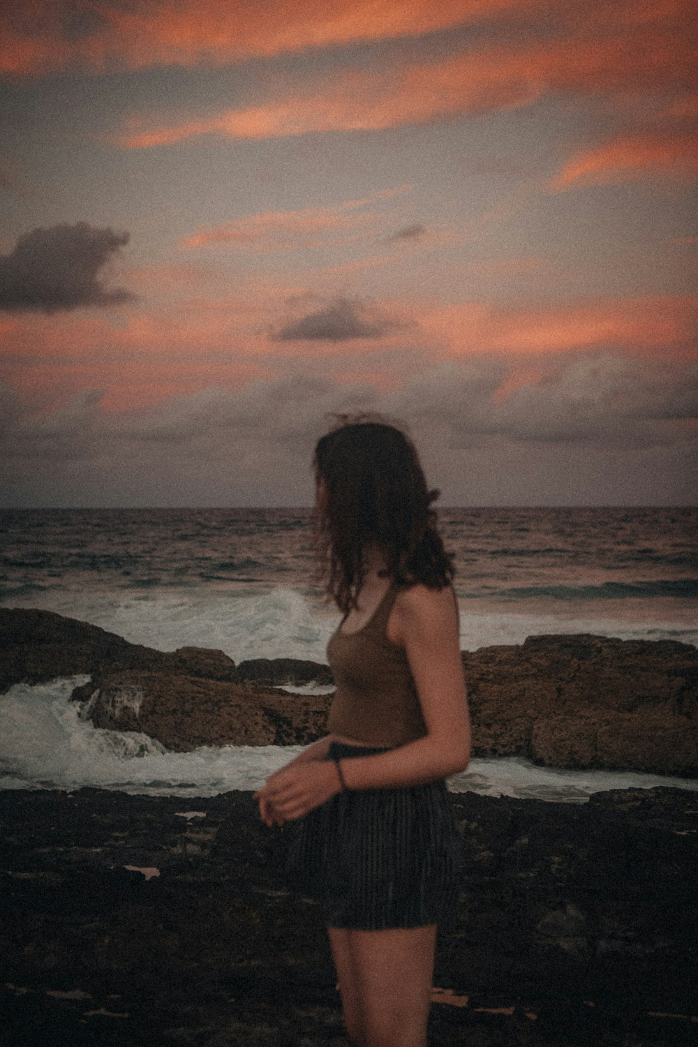 femme en robe grise debout sur la formation rocheuse près de la mer pendant le coucher du soleil