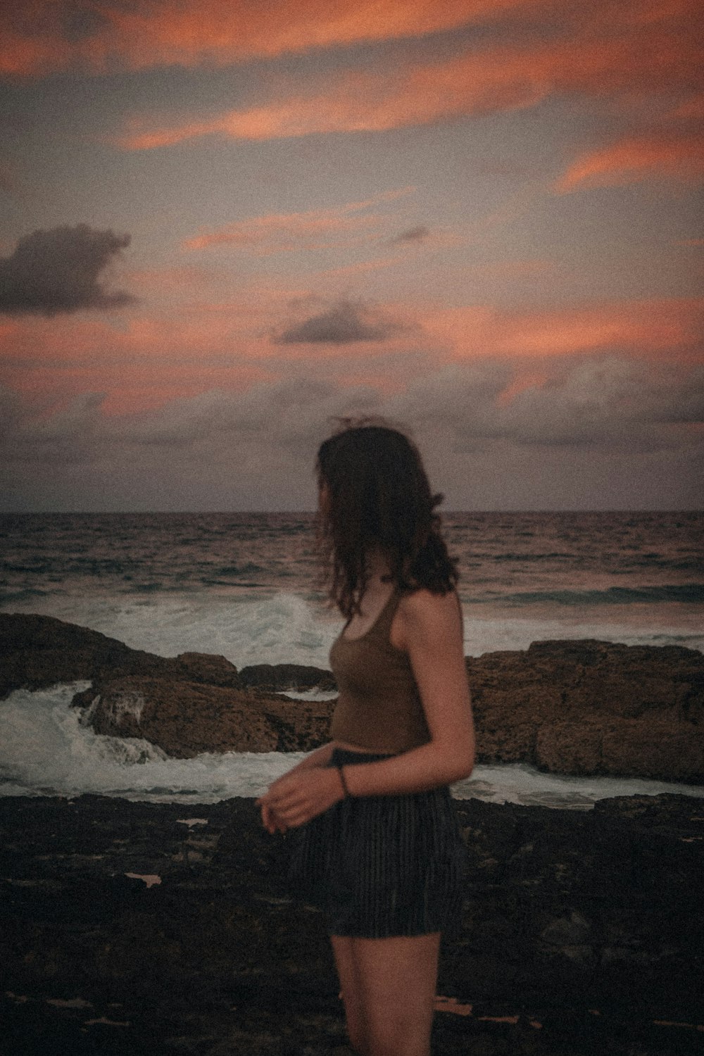Frau in grauem Kleid, die bei Sonnenuntergang auf einer Felsformation in der Nähe des Meeres steht