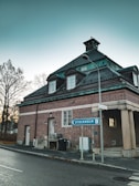 A brick building with a dark green roof sits at a street corner. A road sign points towards Stockholm. Bare trees are visible in the background against a clear sky.