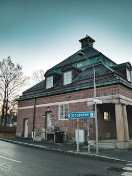 A brick building with a dark green roof sits at a street corner. A road sign points towards Stockholm. Bare trees are visible in the background against a clear sky.