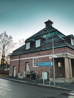 A brick building with a dark green roof sits at a street corner. A road sign points towards Stockholm. Bare trees are visible in the background against a clear sky.