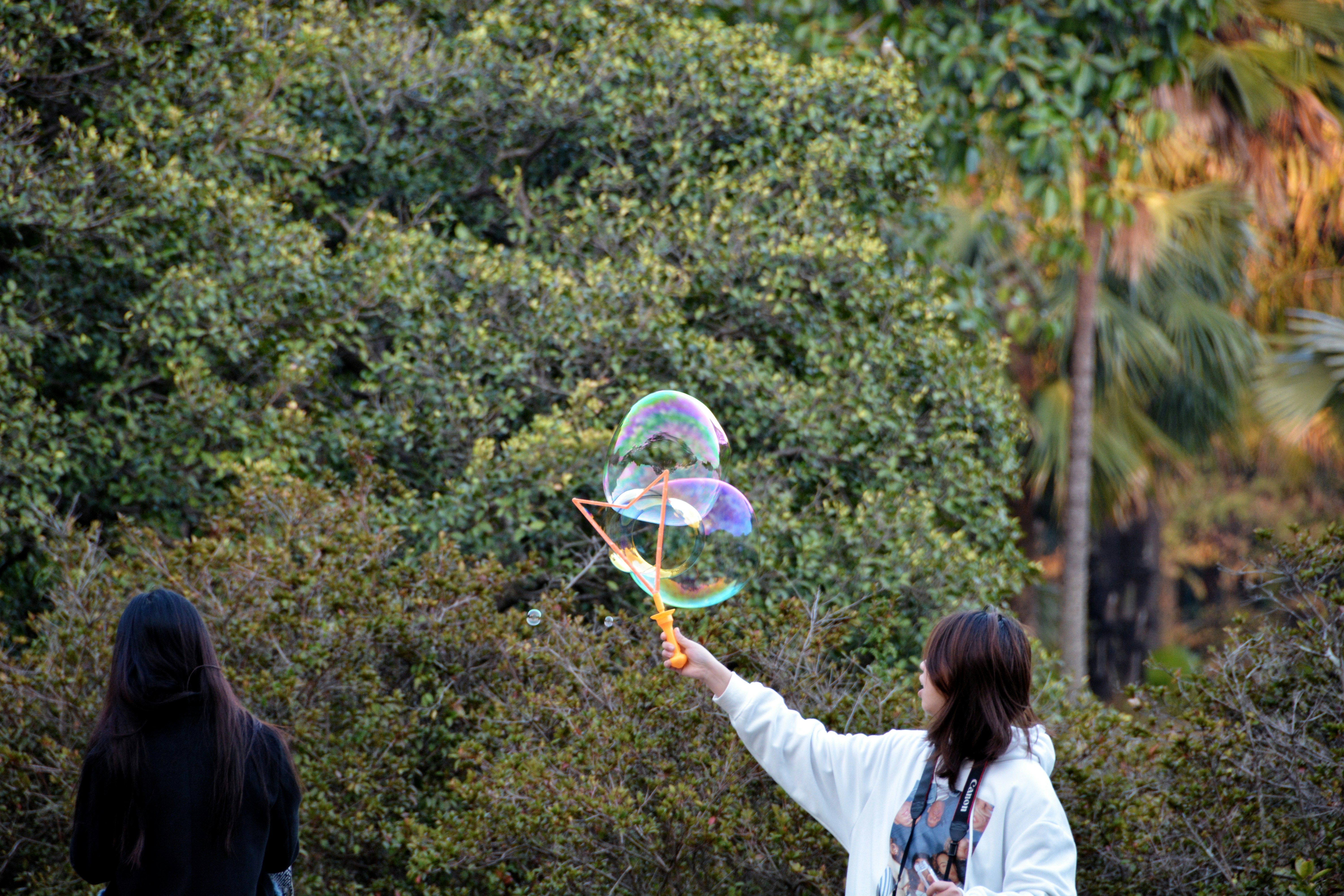 A girl in a white long-sleeve shirt playing with bubbles near green trees during the day