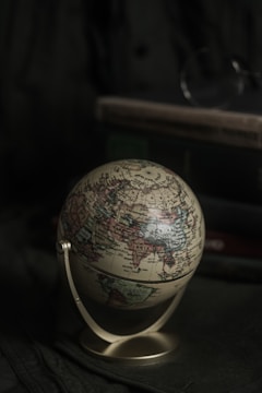 A small globe rests on a surface with a muted green backdrop. The globe displays various countries and continents with intricate details and geographical names. In the background, there is a stack of books partially visible, adding to a scholarly or antique setting.
