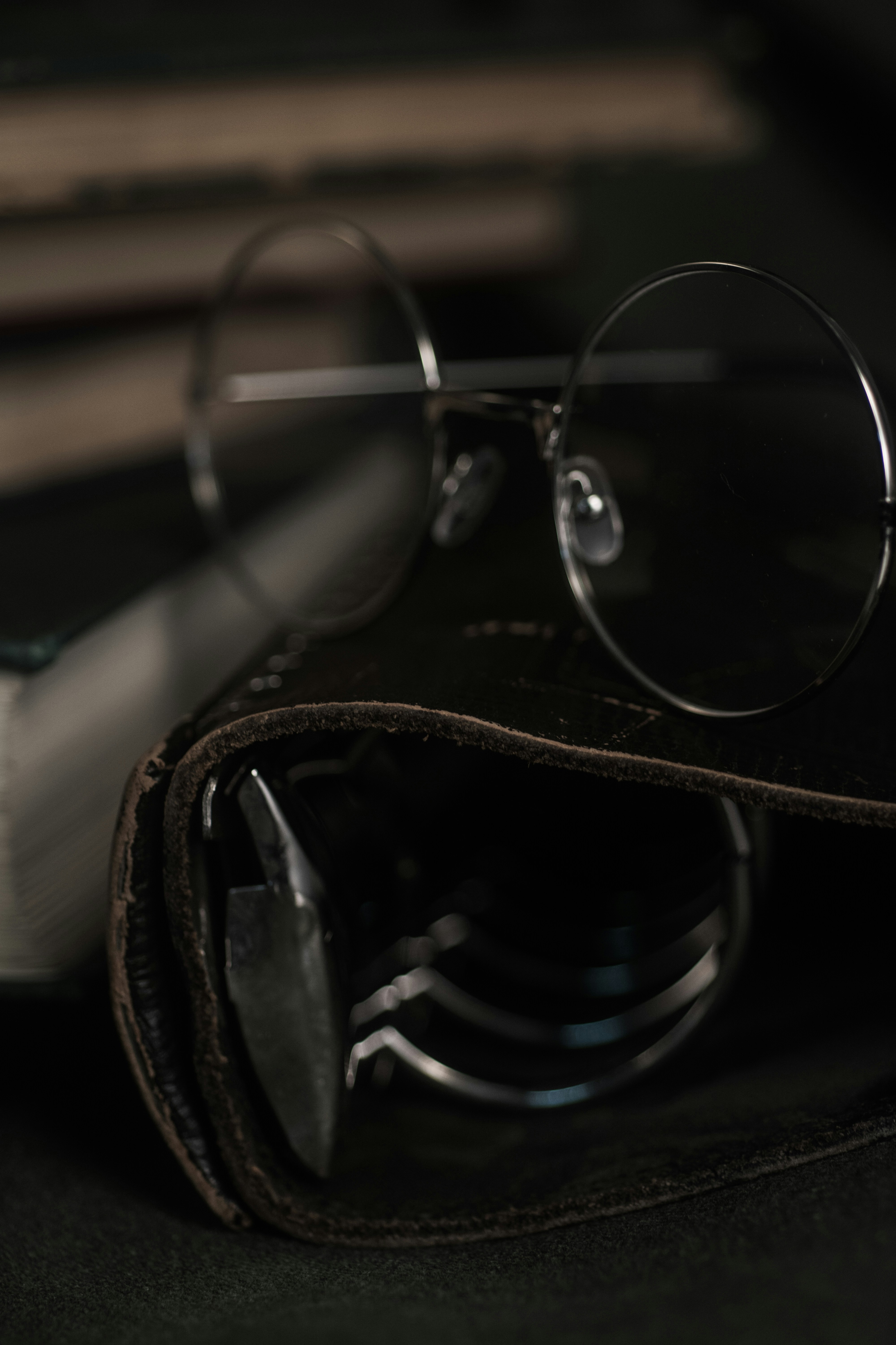 A worn leather journal and reading glasses on a dark wooden table, dim lamp light, deep shadows, intimate and still