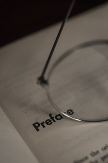 A close-up of a book page featuring the word 'Preface' in bold, partially obscured by a pair of eyeglasses with a thin metallic frame.