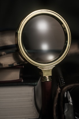 A close-up view of a magnifying glass leaning against a pile of old books with worn edges. The glass has a brass frame and a wooden handle, reflecting light softly in a dimly lit environment.