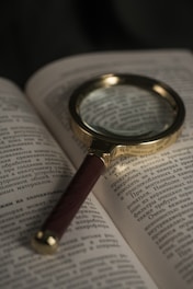 A magnifying glass with a wooden handle rests on an open book filled with text in a foreign language. The lighting is dim, highlighting the glass and the text beneath it.