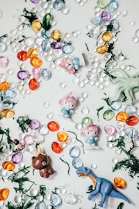 The image features small, colorful toys scattered across a surface. Among them are several miniature figures resembling characters with pink bows and blue outfits, a brown bear with a small yellow object on its head, and a blue dinosaur. Surrounding the figures are vibrant, translucent stones in purple, orange, yellow, blue, and green. There are also small patches of dark green artificial grass interspersed throughout.