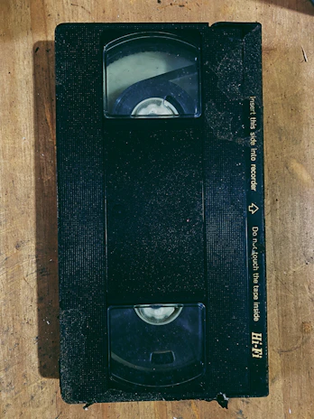 A grainy VHS tape labeled with cryptic symbols resting on a scratched metal surface under shadow.