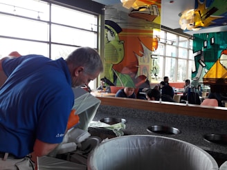 A person wearing a blue shirt and orange gloves is emptying a container into a large trash bin in a cafeteria with colorful comic-style artwork on glass panels. Several people are seated at tables in the background, engaging in conversation.