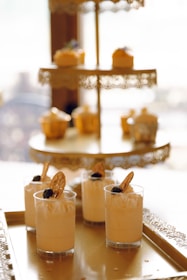 Elegant dessert display featuring a tiered, ornate metal stand. The top tier holds cupcakes with a golden hue, while the bottom tier displays small cups of creamy mousse garnished with blueberries and dried orange slices.