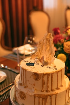 A two-tiered cake decorated with gold accents, dried citrus slices, and elegant drizzles. The top tier features an artistic shard-like decoration and the phrase 'La vita &eacute; bella'. The setting includes a table with fancy dinnerware and a floral arrangement in the background.