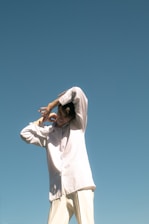 A person wearing a loose, light-colored shirt and trousers stands against a clear blue sky. The individual is smiling and has arms raised above the head in a relaxed, playful posture.