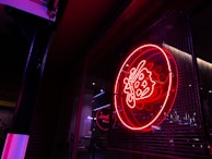 A bright red neon sign with intricate designs, possibly Asian characters, is displayed against a metal grid background. The surrounding area appears dark with some reflections and light from adjacent sources.