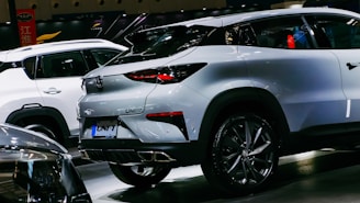 A sleek, modern crossover SUV with a white exterior is showcased in a well-lit indoor setting. The vehicle features stylish, angular rear lights and a dual exhaust design. Other cars are visible in the background, indicating a car show or exhibition environment.