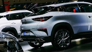 A sleek, modern crossover SUV with a white exterior is showcased in a well-lit indoor setting. The vehicle features stylish, angular rear lights and a dual exhaust design. Other cars are visible in the background, indicating a car show or exhibition environment.