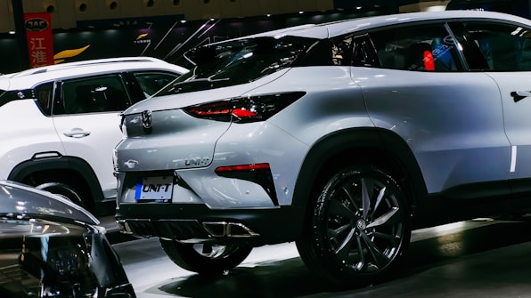 A sleek, modern crossover SUV with a white exterior is showcased in a well-lit indoor setting. The vehicle features stylish, angular rear lights and a dual exhaust design. Other cars are visible in the background, indicating a car show or exhibition environment.