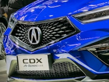 A close-up of the front grille of a blue car, prominently featuring a shiny logo. The car's sleek design includes angular headlights and a stylish honeycomb-patterned grille. The reflection of lights on the car's surface highlights its glossy finish.