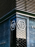 The image features an architectural detail of a modern building with geometric patterns on the facade. The building exterior includes dark stone tiles and a hexagonal glass window design. Prominent logos with the letters 'P' are visible on the glass sections.