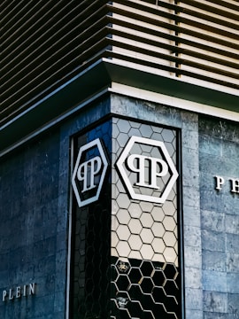 The image features an architectural detail of a modern building with geometric patterns on the facade. The building exterior includes dark stone tiles and a hexagonal glass window design. Prominent logos with the letters 'P' are visible on the glass sections.