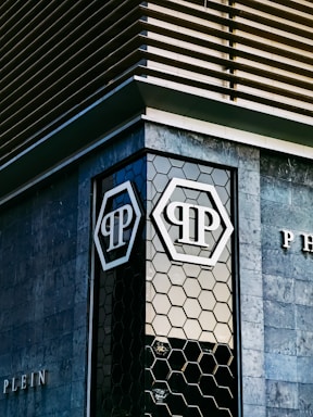 The image features an architectural detail of a modern building with geometric patterns on the facade. The building exterior includes dark stone tiles and a hexagonal glass window design. Prominent logos with the letters 'P' are visible on the glass sections.