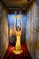 A wooden sculpture is displayed in a small, dimly lit alcove with textured stone walls. The sculpture features a tall, slender figure with elongated arms reaching upwards, holding a golden, lyre-like instrument. The base of the sculpture is adorned with a treble clef symbol. The alcove is draped with a rich, burgundy cloth at the bottom.