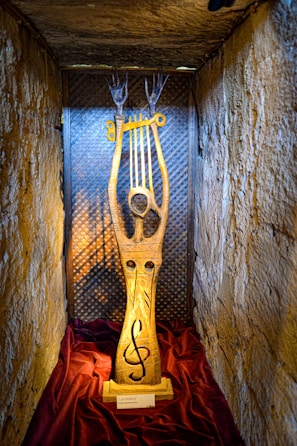 A wooden sculpture is displayed in a small, dimly lit alcove with textured stone walls. The sculpture features a tall, slender figure with elongated arms reaching upwards, holding a golden, lyre-like instrument. The base of the sculpture is adorned with a treble clef symbol. The alcove is draped with a rich, burgundy cloth at the bottom.