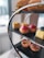 A tiered serving stand displays a selection of pastries and sandwiches, including a sandwich with visible fillings such as cucumber, two pastries with pink icing, a yellow sponge cake, and two small quiches or tarts on the lower tier. The background is softly blurred, suggesting an indoor setting.