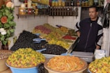 A market stall features a variety of olives and preserved vegetables arranged in large heaps. The vendor stands next to jars of preserved lemons and bottles of oil displayed on shelves. Brightly colored produce, including green and black olives, and pickled vegetables, create a vibrant setup. The overall scene has a rustic, homely market atmosphere.
