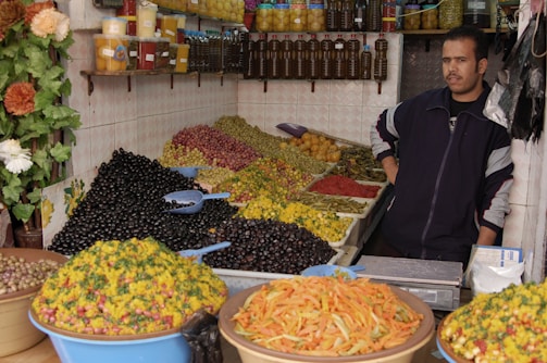 A market stall features a variety of olives and preserved vegetables arranged in large heaps. The vendor stands next to jars of preserved lemons and bottles of oil displayed on shelves. Brightly colored produce, including green and black olives, and pickled vegetables, create a vibrant setup. The overall scene has a rustic, homely market atmosphere.