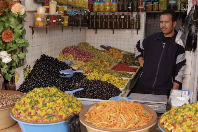 A market stall features a variety of olives and preserved vegetables arranged in large heaps. The vendor stands next to jars of preserved lemons and bottles of oil displayed on shelves. Brightly colored produce, including green and black olives, and pickled vegetables, create a vibrant setup. The overall scene has a rustic, homely market atmosphere.