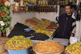 A market stall features a variety of olives and preserved vegetables arranged in large heaps. The vendor stands next to jars of preserved lemons and bottles of oil displayed on shelves. Brightly colored produce, including green and black olives, and pickled vegetables, create a vibrant setup. The overall scene has a rustic, homely market atmosphere.