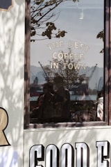 A reflective glass window displays the words 'THE BEST COFFEE IN TOWN'. Outside the window, silhouettes of cranes and what looks like a harbor can be seen under a clear sky. Shadows of tree branches add depth to the scene, and faint reflections of people are visible in the glass.