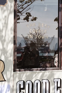 A reflective glass window displays the words 'THE BEST COFFEE IN TOWN'. Outside the window, silhouettes of cranes and what looks like a harbor can be seen under a clear sky. Shadows of tree branches add depth to the scene, and faint reflections of people are visible in the glass.