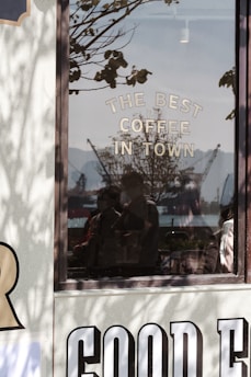 A reflective glass window displays the words 'THE BEST COFFEE IN TOWN'. Outside the window, silhouettes of cranes and what looks like a harbor can be seen under a clear sky. Shadows of tree branches add depth to the scene, and faint reflections of people are visible in the glass.