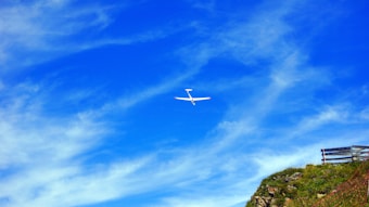 A white glider is soaring through a bright blue sky with scattered white clouds. Below, a grassy hillside with a wooden fence partially visible creates a contrast with the vivid sky.