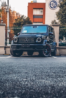 A black luxury SUV is parked in an urban setting. The vehicle is positioned prominently in front of a building with a logo on the top corner. The background includes brick and white facades, trees, and wrought iron fences.