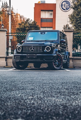 A black luxury SUV is parked in an urban setting. The vehicle is positioned prominently in front of a building with a logo on the top corner. The background includes brick and white facades, trees, and wrought iron fences.
