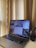 A laptop displaying multiple different applications or windows on its screen is placed on a desk near a set of beige curtains. Next to the laptop on the right is a small black table-top vacuum cleaner.