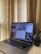 A laptop displaying multiple different applications or windows on its screen is placed on a desk near a set of beige curtains. Next to the laptop on the right is a small black table-top vacuum cleaner.