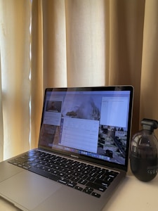 A laptop displaying multiple different applications or windows on its screen is placed on a desk near a set of beige curtains. Next to the laptop on the right is a small black table-top vacuum cleaner.