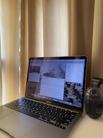 A laptop displaying multiple different applications or windows on its screen is placed on a desk near a set of beige curtains. Next to the laptop on the right is a small black table-top vacuum cleaner.