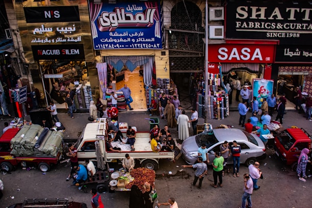 A bustling market scene with people engaged in various activities. Several shops display textiles and fabrics, while vendors sell goods from trucks and stalls on the street. The crowd includes shoppers walking and interacting, contributing to a lively atmosphere.