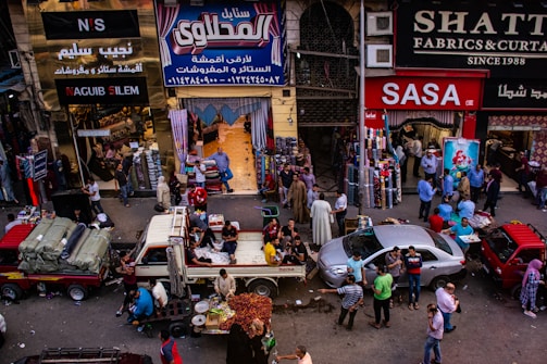 A bustling market scene with people engaged in various activities. Several shops display textiles and fabrics, while vendors sell goods from trucks and stalls on the street. The crowd includes shoppers walking and interacting, contributing to a lively atmosphere.