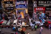 A bustling market scene with people engaged in various activities. Several shops display textiles and fabrics, while vendors sell goods from trucks and stalls on the street. The crowd includes shoppers walking and interacting, contributing to a lively atmosphere.