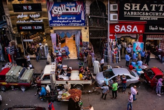 A bustling market scene with people engaged in various activities. Several shops display textiles and fabrics, while vendors sell goods from trucks and stalls on the street. The crowd includes shoppers walking and interacting, contributing to a lively atmosphere.