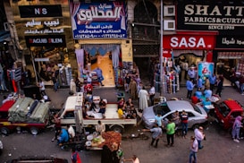 A bustling market scene with people engaged in various activities. Several shops display textiles and fabrics, while vendors sell goods from trucks and stalls on the street. The crowd includes shoppers walking and interacting, contributing to a lively atmosphere.