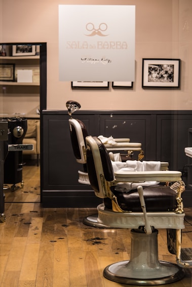 A barbershop interior features vintage-style barber chairs with white and black leather, positioned in front of a wooden floor. The walls are adorned with black-and-white photographs, and a glass panel with the logo and name 'SALA DE BARBA' provides a view into the well-organized space.